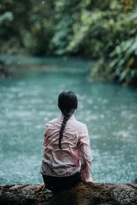 Back view of unrecognizable female traveler sitting on shore of calm celeste river with turquoise water flowing through green rainforest while relaxing and enjoying solitude during summer journey in costa rica