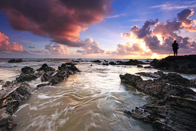 Silhouette man standing on rock formation by sea against sky during sunset