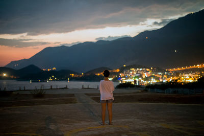 Rear view of woman looking at illuminated city by sea against sky at dusk