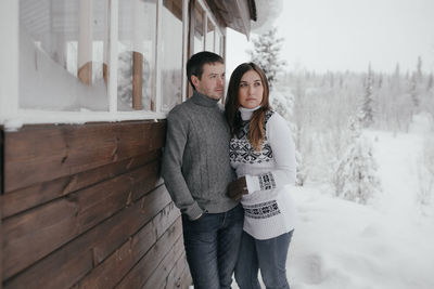 Beautiful couple in love embracing each other, small wooden house and snow in the backgroun