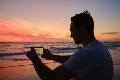 Side view of man photographing sea against sky during sunset