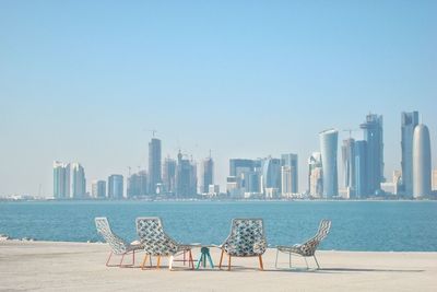 Chairs by sea against buildings in city against clear sky