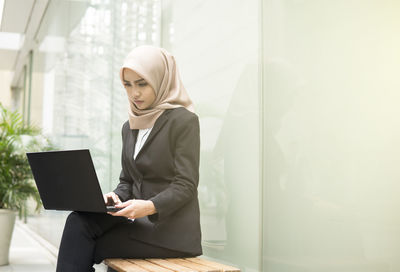 Young businesswoman sitting with laptop on bench at office lobby