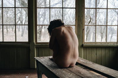 Rear view of shirtless woman sitting on bench in abandoned room
