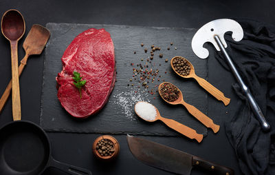 Raw beef tenderloin lies on a cutting board and spices for cooking on a black table, top view