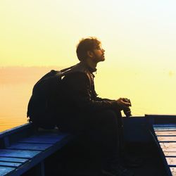 Man with backpack holding camera sitting on boat against sky during sunset