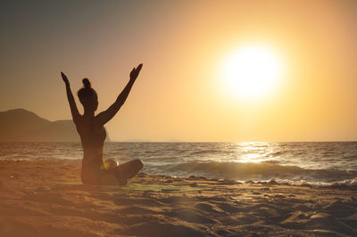 Rear view of woman doing yoga at beach against sky during sunset