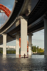 Side view of woman standing on paddle board in river and rowing with paddle under bridge in city