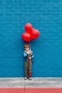 Full length of boy with red balloons standing against blue wall