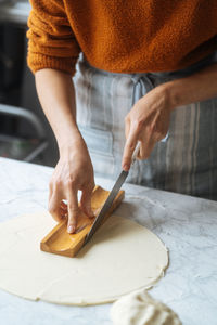 From above crop skilled cook in apron cutting dough with knife using shape on table in kitchen