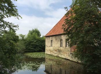 House by river amidst trees and building against sky