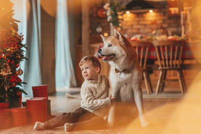 Candid authentic happy little boy in knitted beige sweater hugs dog with bow tie at home on xmas