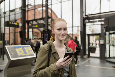 Portrait of young woman holding mobile phone while standing at railroad station