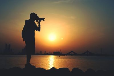 Full length of silhouette man photographing by sea against orange sky