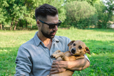 Handsome stylish european man is holding his dog in hands in park on a walk.