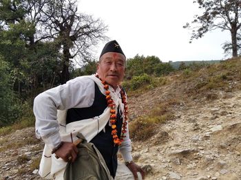 Man wearing cap and floral garland standing on dirt road against sky