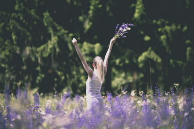 Rear view of woman with arms raised holding flowers in field during sunny day