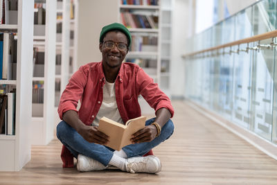 Happy clever black student man with book sitting on floor in university library, looking at camera