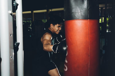 Young focused asian man training boxing performing punches while exercising with heavy punching bag in a modern gym