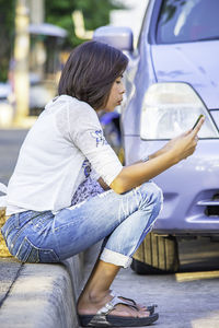 Side view of woman using phone while sitting on seat against car