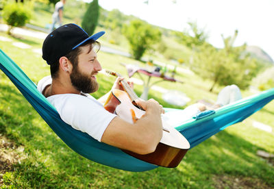 Bearded man with a guitar lying in a hammock smiling against the backdrop of nature and greenery.