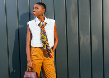 Gorgeous adult african american female entrepreneur with short dark hair in stylish smart casual clothes standing near gray wall with hand in pocket and looking away