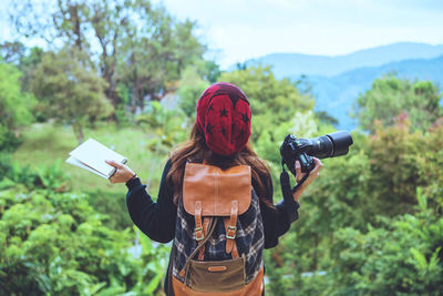 Rear view of young woman with camera standing against mountain