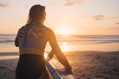 Rear view of woman with surfboard walking towards sea at beach during sunset