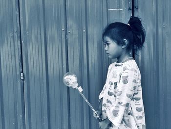 Girl holding toy looking away while standing against wall