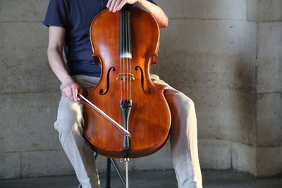 Midsection of man sitting with cello against wall