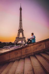 Side view of man looking at eiffel tower while sitting on retaining wall during sunset