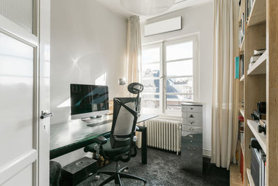 Comfortable armchair placed near table with computer monitor in modern workplace with shelves in contemporary apartment