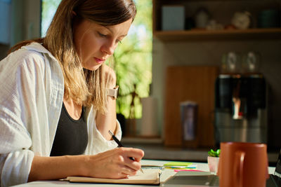 Woman working at home, writing in notebook and using laptop