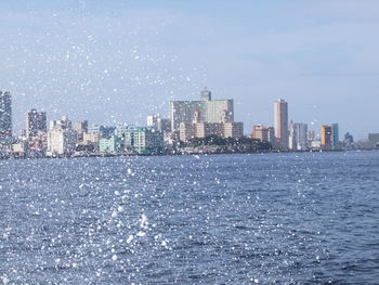 Surf splashing over sea by cityscape against sky