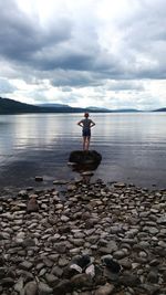 Rear view of girl standing on rock in lake against cloudy sky