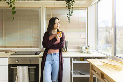 Woman with mug leaning on kitchen counter at home