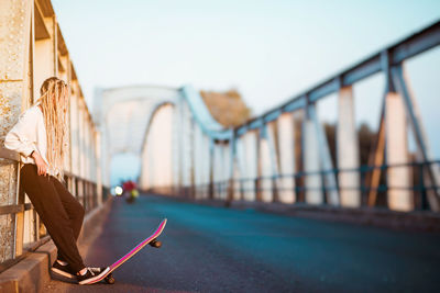 Side view of young woman with skateboard standing on bridge against sky in city during sunset