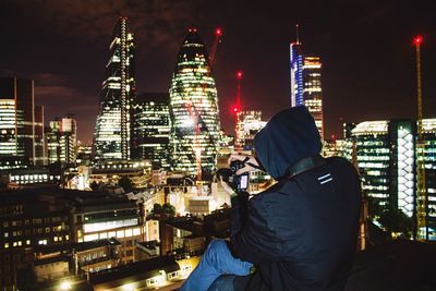 Rear view of man photographing illuminated cityscape against sky