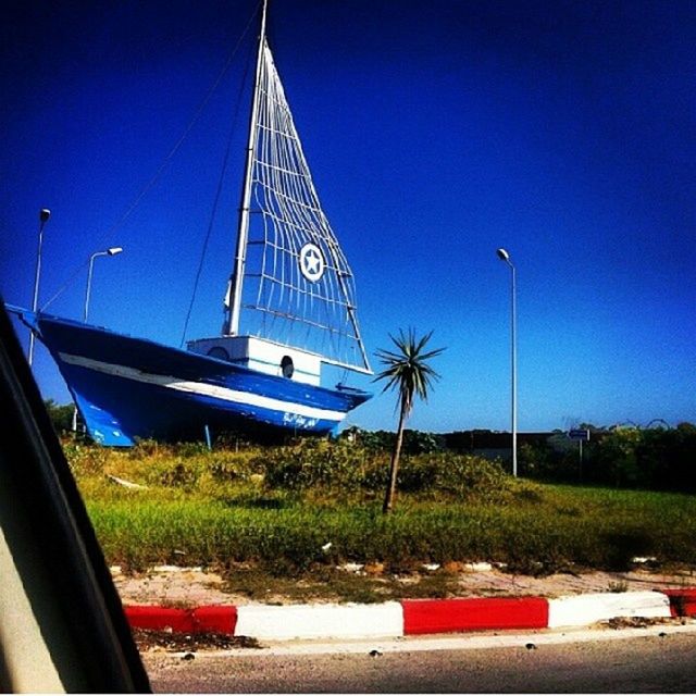 blue, clear sky, transportation, beach, nautical vessel, flag, sky, boat, mode of transport, day, sand, water, shore, outdoors, tree, nature, grass, sea, sunlight, no people