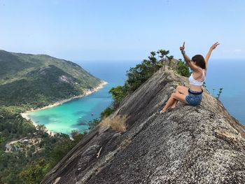 Woman with arms raised sitting on cliff by sea against blue sky