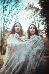 Portrait of sisters wrapped in blanket standing against trees