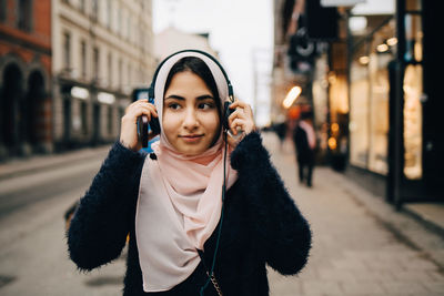 Teenage girl adjusting headphones holding smart phone walking on street in city