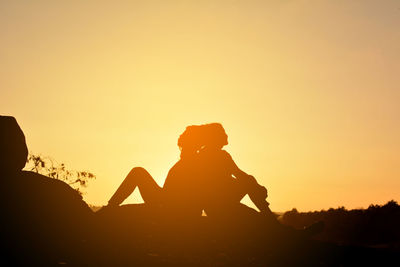 Full length of silhouette friends sitting on rock against clear sky during sunset