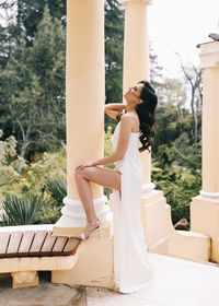 A beautiful brunette lady in an elegant wedding dress poses among the columns in the old city park