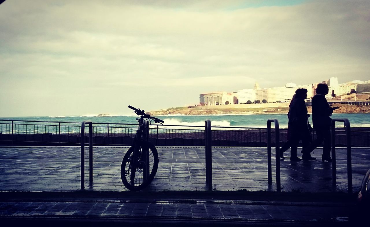 MAN AND BICYCLE ON RAILING BY SEA AGAINST SKY IN CITY