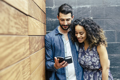 Smiling young couple using mobile phone while standing on wall