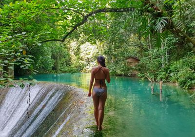 Seductive young woman wearing bikini while standing by waterfall and lake in forest