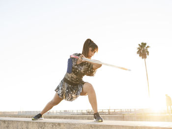 Young female athlete performing martial arts with sword on structure against clear sky