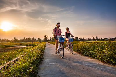 Friends cycling on road against sky during sunset