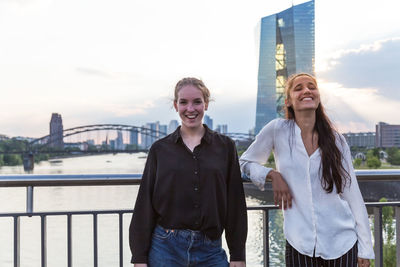 Portrait of cheerful young women standing against railing on bridge in city